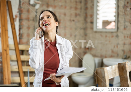 Positive female designer with papers talking on phone in building under renovation Positive female designer with papers talking on phone in building under renovation 118461693