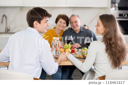 Couple sharing close moment at table, holding hands and locking eyes, while happy parents watching in background at family home. Blessing of parents Couple sharing close moment at table, holding hands and locking eyes, while happy parents watching in background at family home. Blessing of parents 118461817