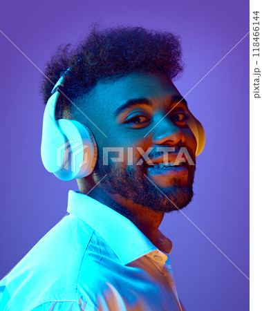 Portrait of young man smiles back at camera with side glance, wearing headphones in vibrant neon light against gradient background. 118466144