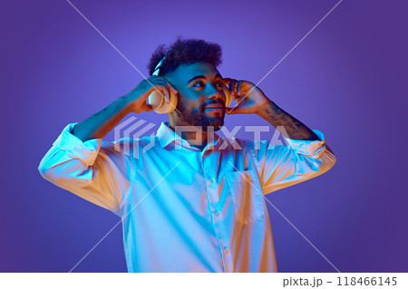 Man looks upward, hands on his white headphones, seemingly lost in rhythm of music. in vibrant neon light against gradient background. 118466145