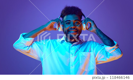 Joyful, relaxed man in white shirt smiles while adjusting his white headphones in vibrant neon light against gradient background. 118466146