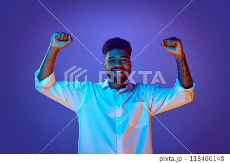 Smiling brightly, man in white shirt raises his fists in triumphant pose in vibrant neon light against gradient background. Smiling brightly, man in white shirt raises his fists in triumphant pose in vibrant neon light against gradient background. 118466148