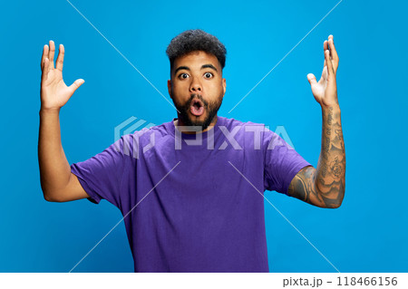 African-American man, wearing purple shirt, looks surprised with his mouth open, raising his hands in playful gesture against blue background. 118466156