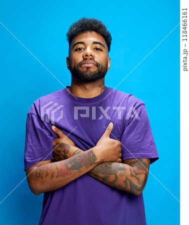 Young man in purple shirt stands confidently, crossing his tattooed arms, looking at camera with confidence against blue background. Young man in purple shirt stands confidently, crossing his tattooed arms, looking at camera with confidence against blue background. 118466161