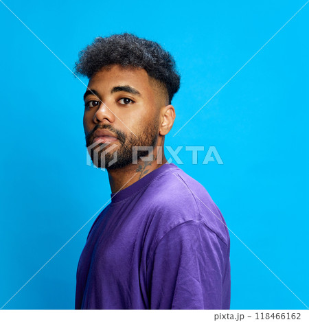 Young African-American man in stylish purple shirt glances at camera with serious expression against blue background. Young African-American man in stylish purple shirt glances at camera with serious expression against blue background. 118466162