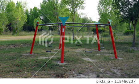 An empty playground features a large swing set with multiple swings, set in a peaceful park. Lush green trees surround the area, creating a calm atmosphere. An empty playground features a large swing set with multiple swings, set in a peaceful park. Lush green trees surround the area, creating a calm atmosphere. 118466237