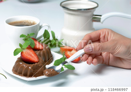 Woman eats dessert. Chocolate fudge garnished with half a strawberry with petals. Garnished with sprigs of mint and strawberries. Near a cup of coffee. On the white surface of the table. Woman eats dessert. Chocolate fudge garnished with half a strawberry with petals. Garnished with sprigs of mint and strawberries. Near a cup of coffee. On the white surface of the table. 118466997