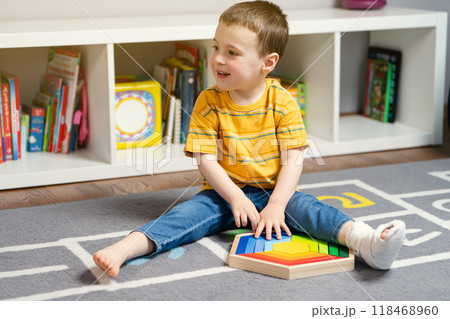 Toddler boy with a bandage or cast on his leg plays with wooden blocks. Fracture of a foot and finger in children. Human healthcare and medicine concept Toddler boy with a bandage or cast on his leg plays with wooden blocks. Fracture of a foot and finger in children. Human healthcare and medicine concept 118468960