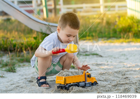 Little cute caucasian toddler boy of three and a half years old plays in the sandbox on a sunny summer day. Outdoor creative activities for kids. Toy dump truck and shovel. Little cute caucasian toddler boy of three and a half years old plays in the sandbox on a sunny summer day. Outdoor creative activities for kids. Toy dump truck and shovel. 118469039