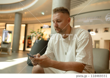 Profile of a happy guy in stylish casual clothes using a smart phone sitting on a soft chair at shopping mall 118469331