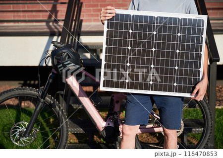 Close up view of person holding solar panel for charging, stands in front of brick building. Next to him pink electric mountain bike with black helmet resting on handlebars. Cropped view. Close up view of person holding solar panel for charging, stands in front of brick building. Next to him pink electric mountain bike with black helmet resting on handlebars. Cropped view. 118470853