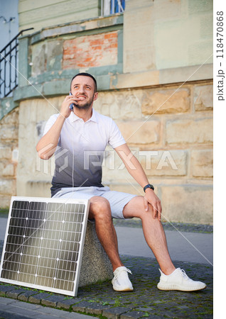 Handsome man using smartphone connected to photovoltaic solar panel. Integration of sustainable renewable energy into everyday life, demonstrating practical use of solar power for charging devices. 118470868