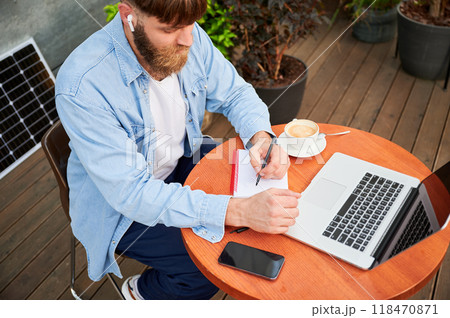 Top view of bearded man works at round wooden table with laptop, notebook, and cup of coffee. Guy wears earbuds, surrounded by greenery and solar panel, highlighting eco-friendly, productive workspace 118470871