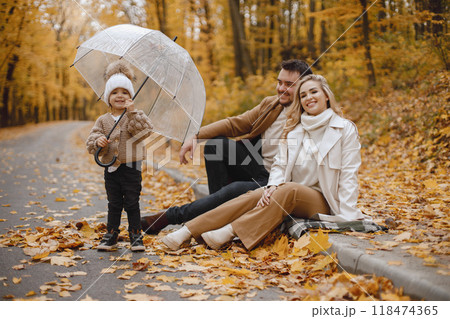 Young man, woman and their daughter sitting outside in autumn forest. Blond mother and brunette father hugging. Little girl holding a transparent umbrella. Young man, woman and their daughter sitting outside in autumn forest. Blond mother and brunette father hugging. Little girl holding a transparent umbrella. 118474365