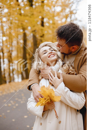 Young man and woman walking outside wearing beige coats. Blond woman and brunette man in autumn forest holding a yellow leaves. Romantic couple hugging. Young man and woman walking outside wearing beige coats. Blond woman and brunette man in autumn forest holding a yellow leaves. Romantic couple hugging. 118474366