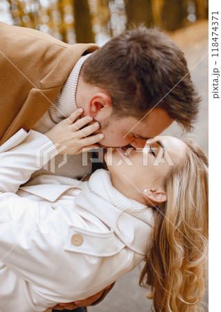 Young man and woman walking outside wearing beige coats. Blond woman and brunette man in autumn forest. Romantic couple kissing. 118474371