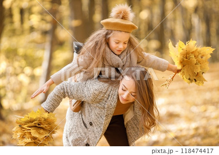 Young woman and little girl in autumn forest. Woman holding her daughter piggyback. Girl wearing fashion clothes and holding yellow leaves. 118474417