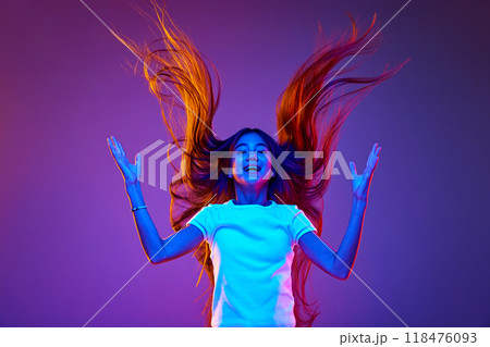 Portrait of joyful girl with long, flowing hair flips it dramatically in air against gradient violet backdrop. Energy radiates through the dynamic colors 118476093