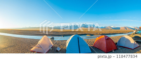Four tents stand on rocky ground next to a quiet river and are surrounded by majestic mountains. There is one car parked near the camp, adding a touch of adventure to this picturesque landscape. 118477834