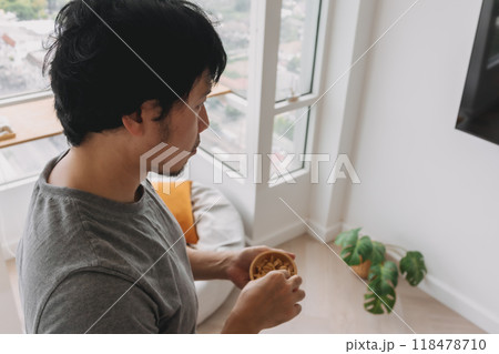 Man standing and eating peanuts snack while watching television in the room. Man standing and eating peanuts snack while watching television in the room. 118478710