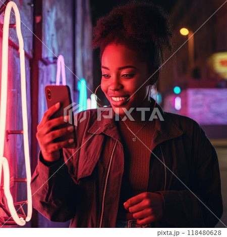 A woman stands in front of a neon-lit wall at night, smiling at her phone, her trendy outfit and the urban energy contrasting her calm, joyful expression. 118480628