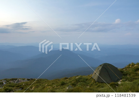 Summer camping in mountains at dawn. Tourist tent on round grassy hill on distant misty blue mountains range under pink sky before sunrise or sunset. Tourism, hiking and beauty of nature concept. Summer camping in mountains at dawn. Tourist tent on round grassy hill on distant misty blue mountains range under pink sky before sunrise or sunset. Tourism, hiking and beauty of nature concept. 118481539