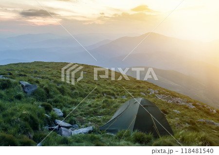 Summer camping in mountains at dawn. Tourist tent on round grassy hill on distant misty blue mountains range under pink sky before sunrise or sunset. Tourism, hiking and beauty of nature concept. Summer camping in mountains at dawn. Tourist tent on round grassy hill on distant misty blue mountains range under pink sky before sunrise or sunset. Tourism, hiking and beauty of nature concept. 118481540