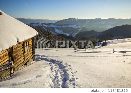 Human footprint path in white deep snow leading to small old wooden forsaken shepherd hut in mountain valley, spruce forest, woody dark hills, bright sun on clear blue sky copy space background. Human footprint path in white deep snow leading to small old wooden forsaken shepherd hut in mountain valley, spruce forest, woody dark hills, bright sun on clear blue sky copy space background. 118481836