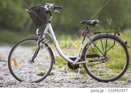 Close-up of new modern white bicycle with black wheels and basket standing on side stand on lit by sun pebbles on blurred green summer bokeh background. Comfortable transport for traveling concept. Close-up of new modern white bicycle with black wheels and basket standing on side stand on lit by sun pebbles on blurred green summer bokeh background. Comfortable transport for traveling concept. 118481957