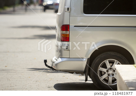 Close-up side view detail of silver passenger medium size luxury minibus van with tow-bar parked on summer sunny city street pavement with blurred silhouettes of pedestrians and cars in background. Close-up side view detail of silver passenger medium size luxury minibus van with tow-bar parked on summer sunny city street pavement with blurred silhouettes of pedestrians and cars in background. 118481977