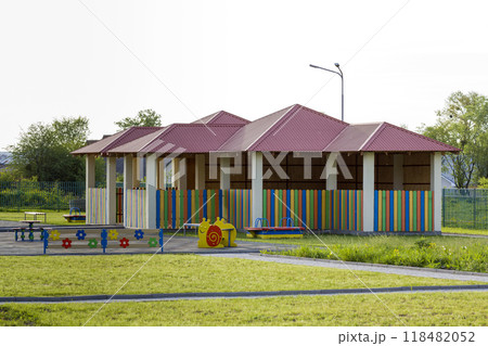 Beautiful playground in kindergarten with bright new alcove with red tile roof and multicolored low fence, green lawn, swings, slides and sandbox. Perfect place for children activities outdoors. 118482052