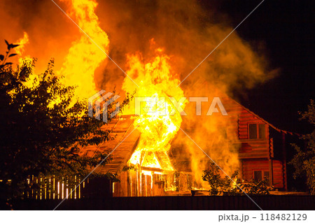 Burning wooden house at night. Bright orange flames and dense smoke from under the tiled roof on dark sky, trees silhouettes and residential neighbor cottage background. Disaster and danger concept. Burning wooden house at night. Bright orange flames and dense smoke from under the tiled roof on dark sky, trees silhouettes and residential neighbor cottage background. Disaster and danger concept. 118482129