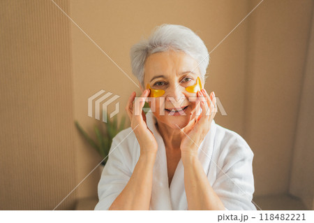 Eye patch beauty aging treatment. Mature adult old woman with under eye collagen pads. Senior woman with patches under eyes gets facial treatment. Senior lady in robe relaxing in bathroom at home Eye patch beauty aging treatment. Mature adult old woman with under eye collagen pads. Senior woman with patches under eyes gets facial treatment. Senior lady in robe relaxing in bathroom at home 118482221