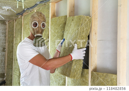 Young worker in gas mask insulating rock wool insulation staff in wooden frame for future house walls for cold and heat barrier. Comfortable warm home, economy, construction and renovation concept. Young worker in gas mask insulating rock wool insulation staff in wooden frame for future house walls for cold and heat barrier. Comfortable warm home, economy, construction and renovation concept. 118482856