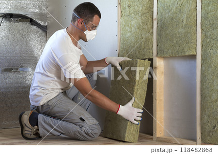 Worker in protective goggles and respirator insulating rock wool insulation in wooden frame for future house walls for cold barrier. Comfortable warm home, economy, construction and renovation concept 118482868