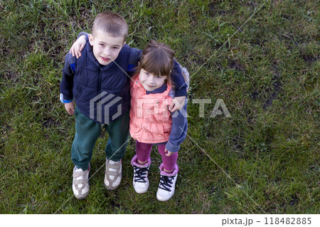 View from above of two cute children in casual clothes boy and girl standing on green grass, hugging each other over shoulders, looking up and shyly smiling. Happy childhood and friendship concept. 118482885