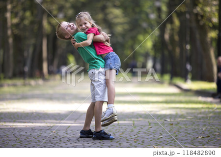 Two cute young funny smiling children, girl and boy, brother holding sister in his arms, having fun on blurred bright sunny park alley green trees bokeh background. Loving siblings relations concept. Two cute young funny smiling children, girl and boy, brother holding sister in his arms, having fun on blurred bright sunny park alley green trees bokeh background. Loving siblings relations concept. 118482890