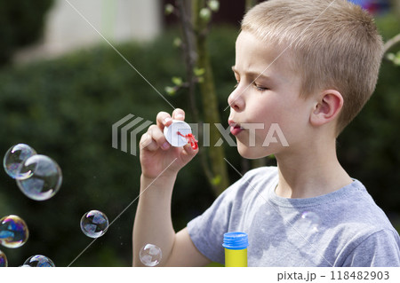 Profile of cute handsome small blond child boy with funny serious expression blowing colorful transparent soap bubbles outdoors on blurred green summer background. Joy of careless childhood concept. 118482903