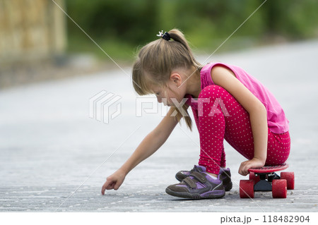 Pretty young long-haired blond child girl in casual pink clothing sitting on skateboard on paved suburb street on blurred sunny summer green background. Children activities, games and fun concept. 118482904