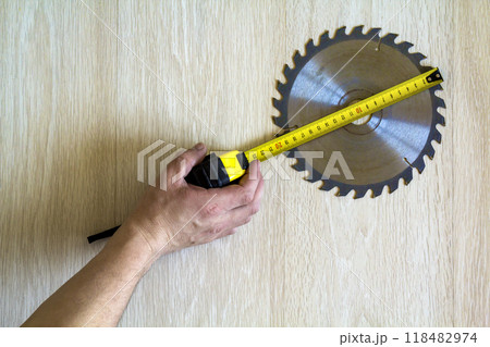 Close up of circular saw blade for wood work and worker hand with measuring tape on wooden background. Top view. Close up of circular saw blade for wood work and worker hand with measuring tape on wooden background. Top view. 118482974