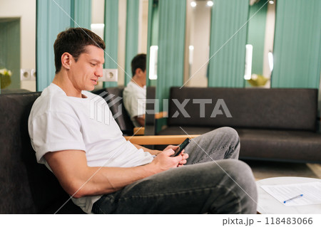 Happy muscular man traveler in white clothes relaxing in hotel lobby using smartphone watching online video or browsing internet. Male tourist resting with phone in lounge area at luxury resort. Happy muscular man traveler in white clothes relaxing in hotel lobby using smartphone watching online video or browsing internet. Male tourist resting with phone in lounge area at luxury resort. 118483066