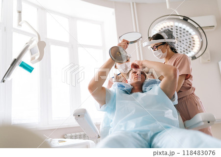 Low-angle portrait of male patient with hairloss problem holding mirror, having consultation with trichologist, marking with pencil new hairline on head before hair transplantation surgery in clinic. Low-angle portrait of male patient with hairloss problem holding mirror, having consultation with trichologist, marking with pencil new hairline on head before hair transplantation surgery in clinic. 118483076