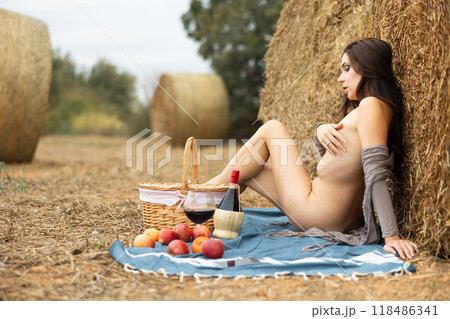 Naked woman in light cape poses on picnic near hay bales in summer field 118486341