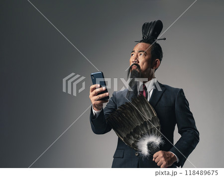 三国志に登場する諸葛孔明のような中国歴史上の軍師がビジネススーツを着てスマホを使うシーン 118489675