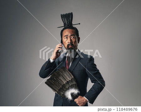 三国志に登場する諸葛孔明のような中国歴史上の軍師がビジネススーツを着てスマホで通話するシーン 118489676