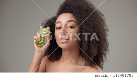 Beauty, vitamins and healthcare concept - beautiful African American woman with curly afro hairstyle and clean, healthy skin holding kiwi halves in her hands posing and looking at the camera 118491795