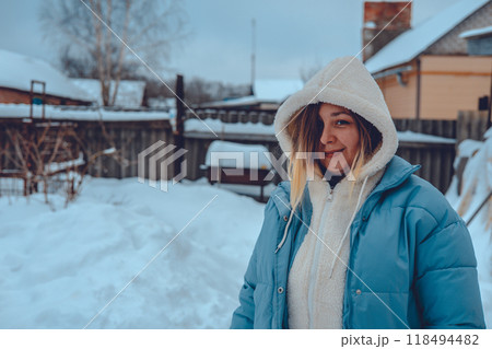 A sweet woman stands against the backdrop of a snowy backyard in winter and smiles tenderly while looking at the camera. Copy space. Portrait of a woman against the backdrop of a snowy winter yard. A sweet woman stands against the backdrop of a snowy backyard in winter and smiles tenderly while looking at the camera. Copy space. Portrait of a woman against the backdrop of a snowy winter yard. 118494482