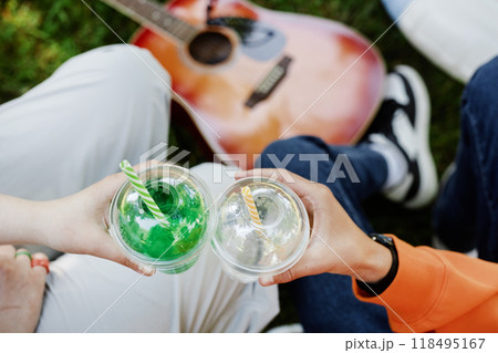 Close up on hands of teenagers holding plastic cups with refreshing drinks while clinking glasses and enjoying picnic in park on sunny summer day, copy space 118495167