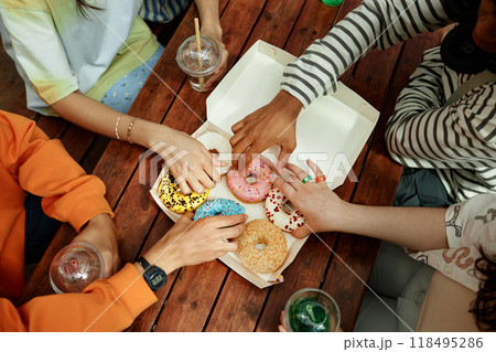 High angle shot of unrecognizable teenagers reaching for delicious sprinkled donut from takeaway box placed on cafe table while partying outdoors High angle shot of unrecognizable teenagers reaching for delicious sprinkled donut from takeaway box placed on cafe table while partying outdoors 118495286