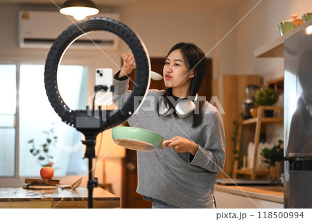 Cheerful young female food blogger recording video in kitchen with a smartphone on a tripod stand with a ring light Cheerful young female food blogger recording video in kitchen with a smartphone on a tripod stand with a ring light 118500994